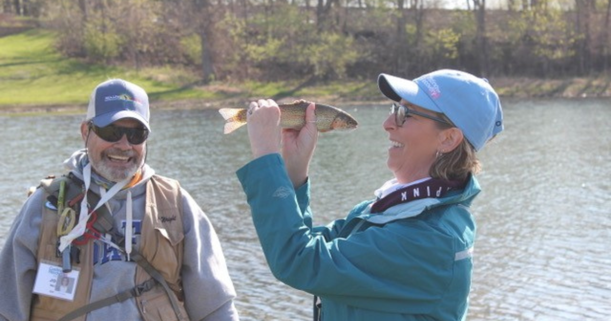 Woman next to a lake holding a fish up to her face and smiling while a smiling man watches