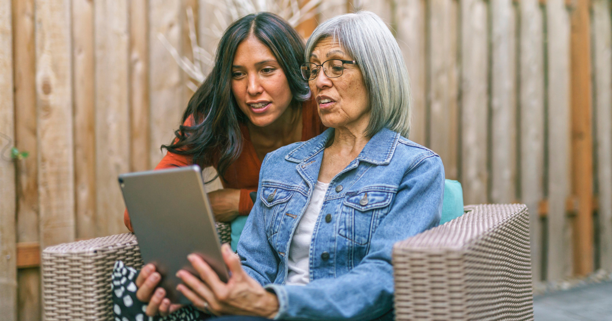 A retired mixed race senior woman sits on outdoor furniture on the back patio of her home. She is holding a tablet computer and her adult daughter is teaching her how to use it.