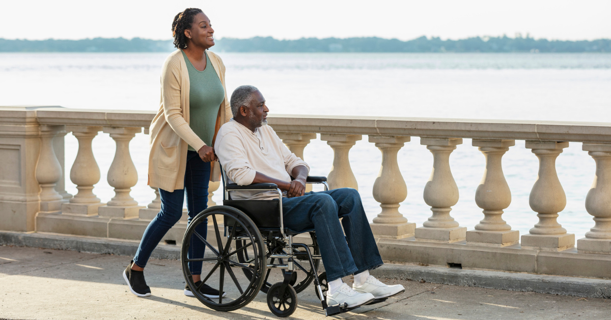 A caregiver pushing a senior African-American man in a wheelchair along a city waterfront. 