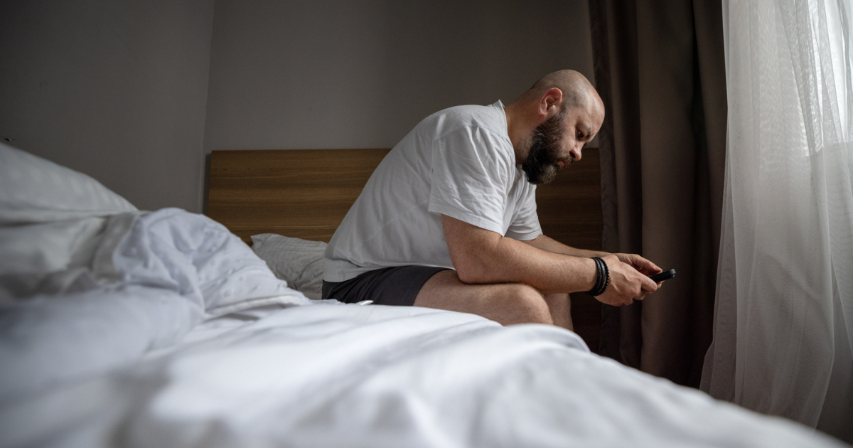  Bearded Man Sitting On Bed In Bedroom Looking At Phone
