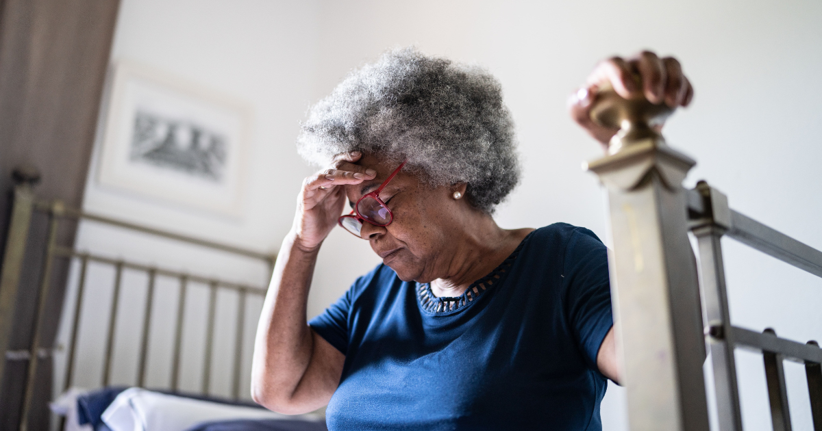 A picture of an older woman sitting on her bed holding her head