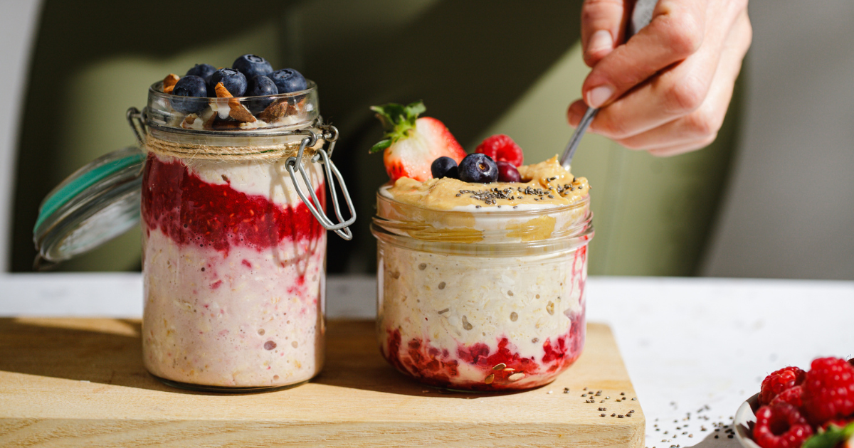 Two glass jars filled with layered overnight oats topped with blueberries, raspberries, strawberry, nuts, chia seeds, and peanut butter. A hand is spooning oats from one jar placed on a wooden board in natural light.
