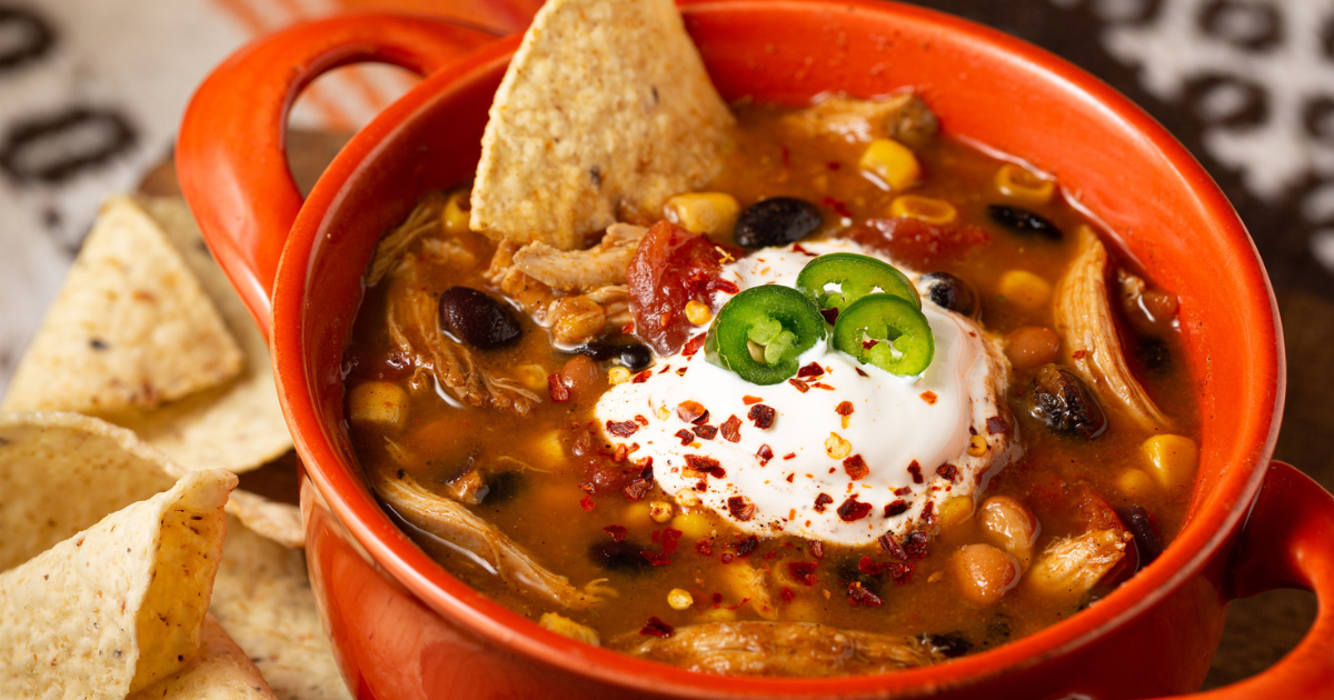 Close-up of a bowl of chicken taco soup topped with sour cream and Serrano peppers with tortilla chips in an orange colored bowl