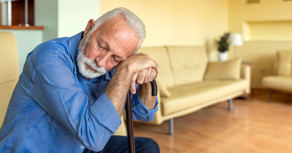 Older man sitting down leaning his head on his cane