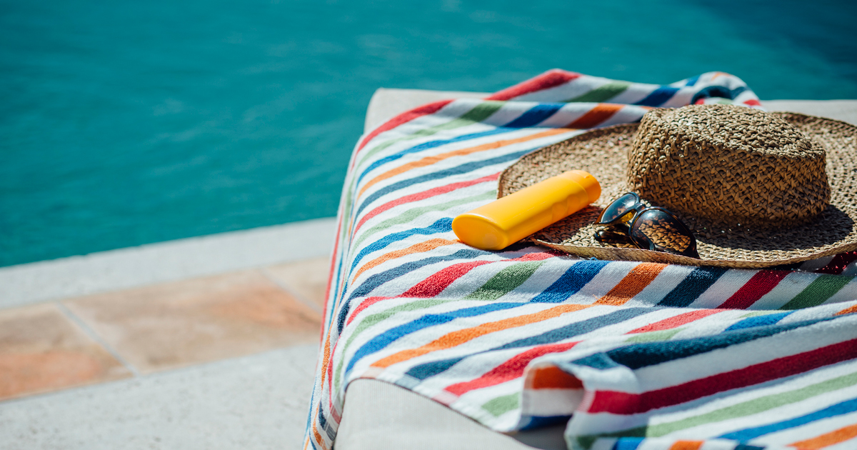 A sun hat, sunglasses, and a bottle of sunscreen on top of a towel on a chair by a pool