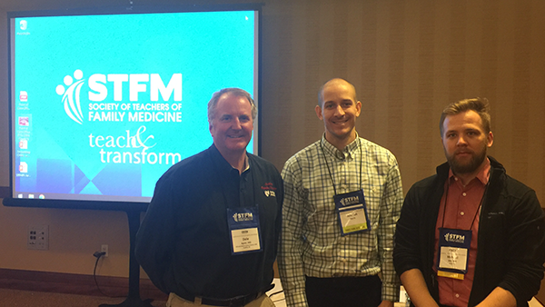 Three men standing in front of a projected image of STFM