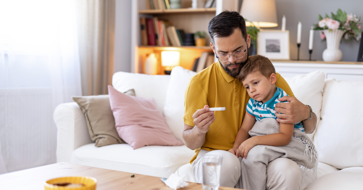 A concerned dad holds a thermometer while his sick child rests on his lap on a white couch