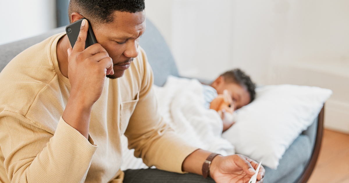 Father on the phone holding a thermometer with his sick child lying on the couch