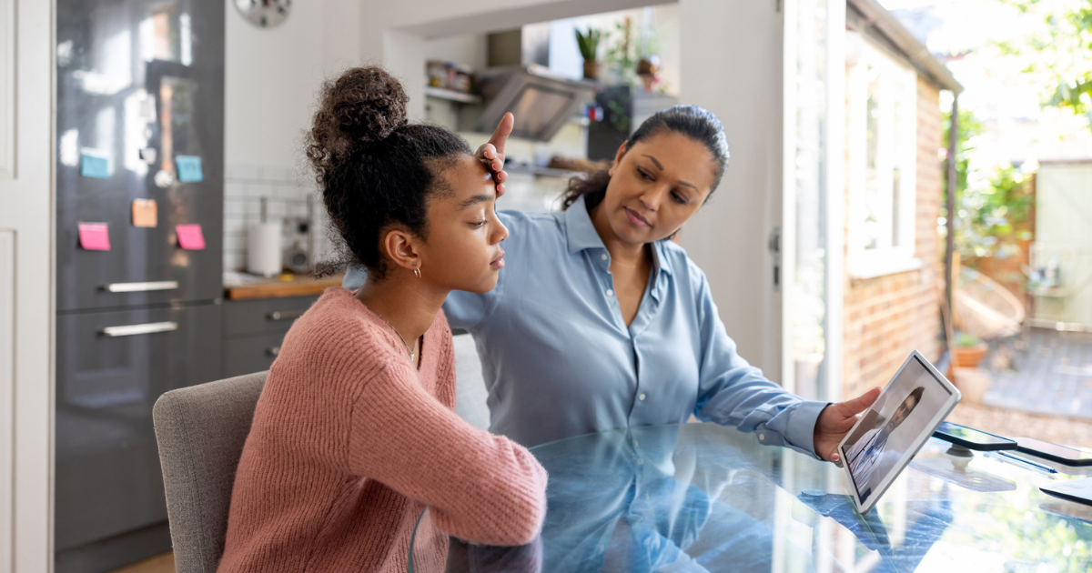 Mother feeling her teen daughter's forehead while on a telehealth appointment with a doctor on a tablet