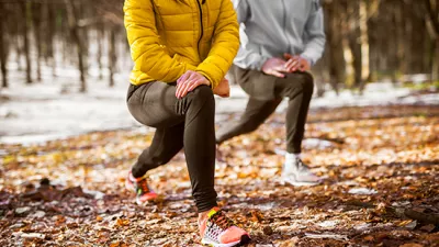 Women wearing running gear stretching in a snowy wooded area. 