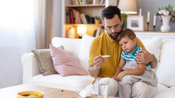 A concerned dad holds a thermometer while his sick child rests on his lap on a white couch
