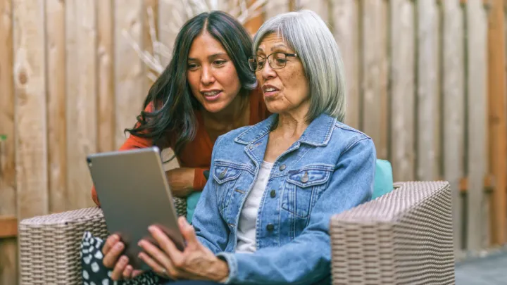 A retired mixed race senior woman sits on outdoor furniture on the back patio of her home. She is holding a tablet computer and her adult daughter is teaching her how to use it.