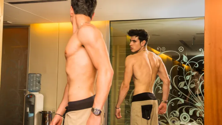 Young Shirtless Handsome Muscular Man Admiring His Muscles in Gym Mirror