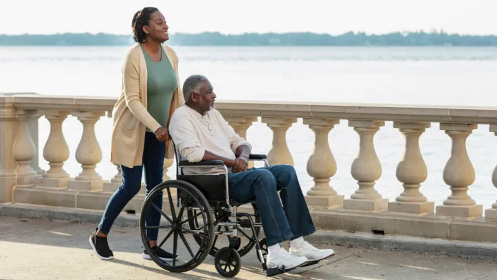 A caregiver pushing a senior African-American man in a wheelchair along a city waterfront. 