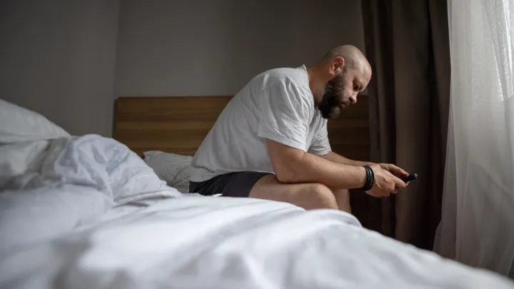  Bearded Man Sitting On Bed In Bedroom Looking At Phone