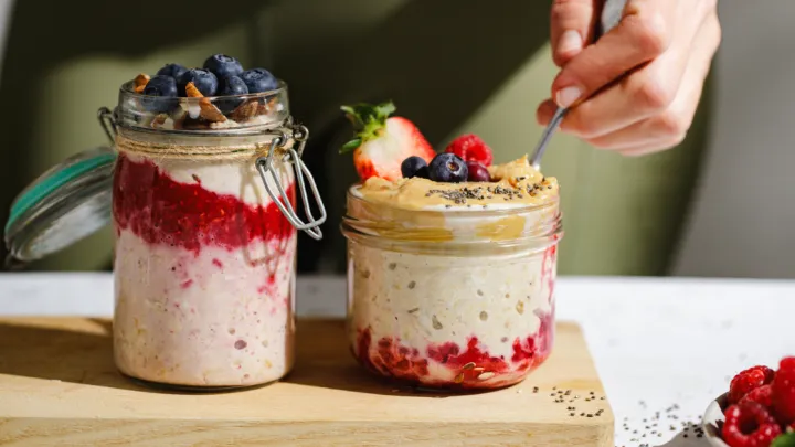 Two glass jars filled with layered overnight oats topped with blueberries, raspberries, strawberry, nuts, chia seeds, and peanut butter. A hand is spooning oats from one jar placed on a wooden board in natural light.