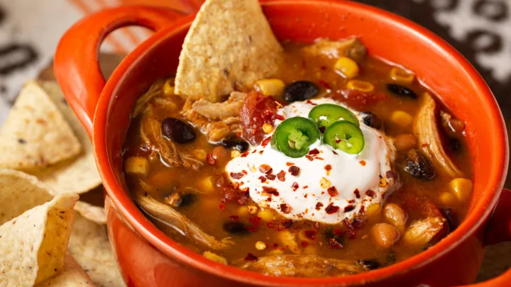 Close-up of a bowl of chicken taco soup topped with sour cream and Serrano peppers with tortilla chips in an orange colored bowl