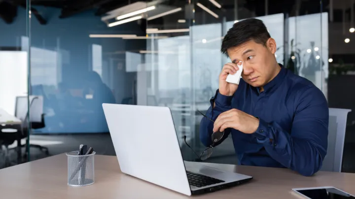 Man sitting at desk, wiping eyes with a tissue