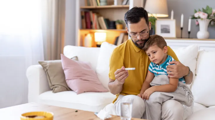 A concerned dad holds a thermometer while his sick child rests on his lap on a white couch