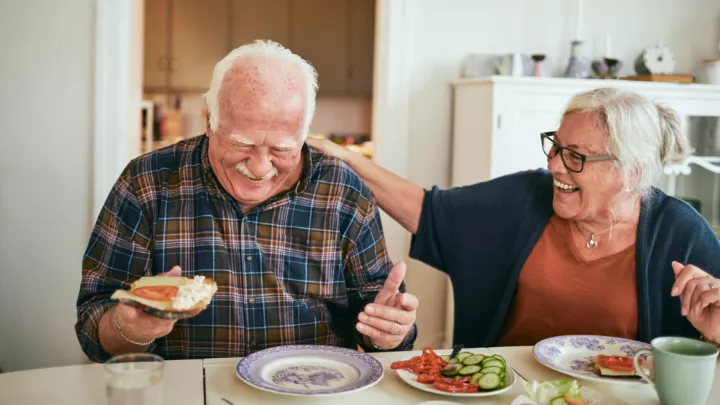 An older couple eating breakfast and laughing at the kitchen table
