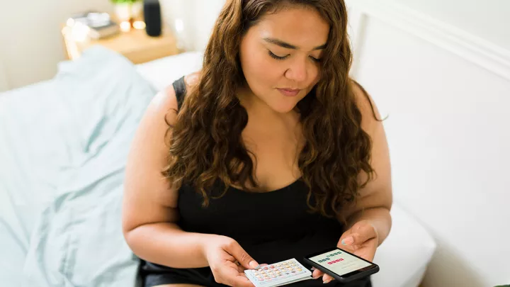 Young woman sitting on her bed, holding birth control pills and her cell phone