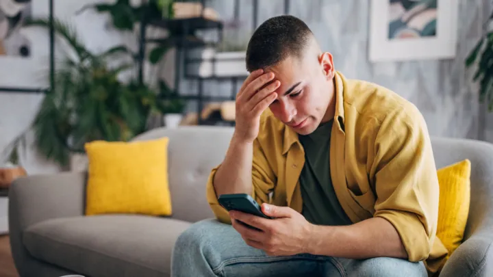 An individual is looking worried while browsing his smartphone , seated with a coffee cup on the table in a peaceful home setting