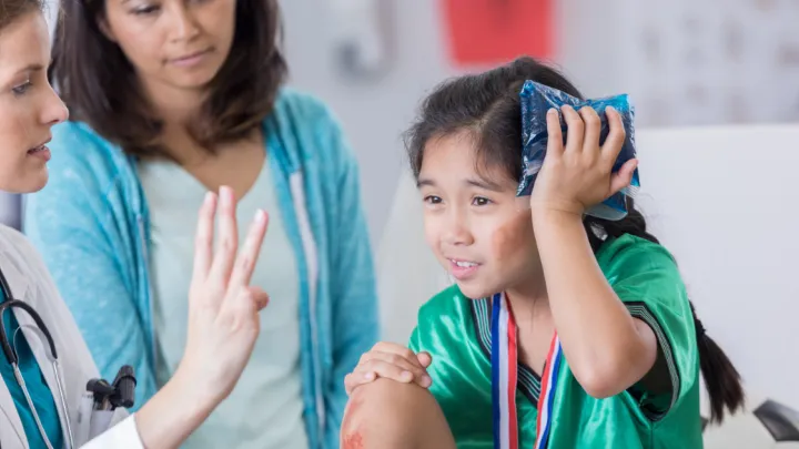 Young girl in a soccer uniform holding an ice pack against her head while a doctor holds up three fingers
