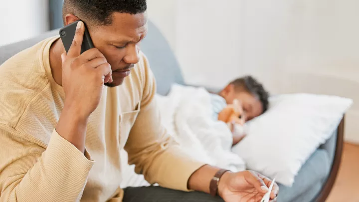 Father on the phone holding a thermometer with his sick child lying on the couch