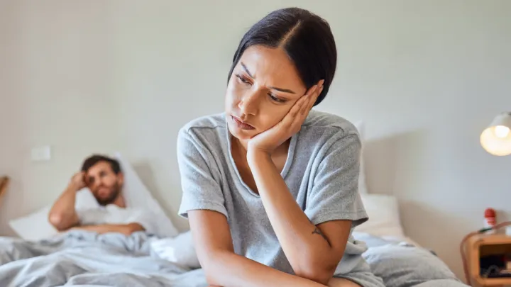 Sad woman sitting on bed with upset man in background